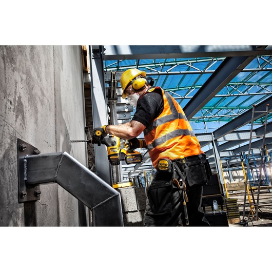 A construction worker, wearing a safety helmet, ear protection, face mask, and orange reflective vest, is using a DEWALT cordless drill to work on a concrete wall at an industrial job site with metal structures and scaffolding.