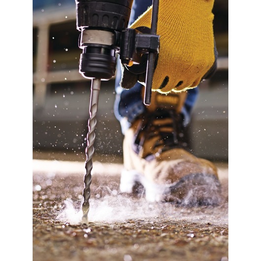 Close-up of a person wearing a yellow work glove using a DEWALT drill with a masonry bit to drill into a concrete floor, with dust scattering around the drill point. The person is also wearing sturdy work boots.