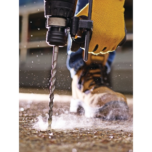 Close-up of a gloved hand using a drill bit on concrete, with dust and work boot visible.