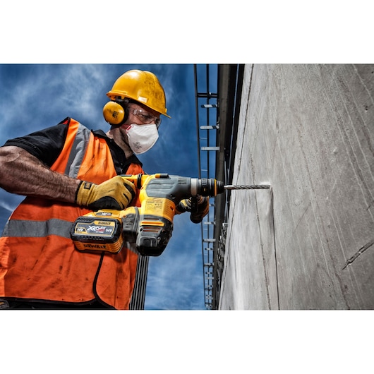 Construction worker drills into concrete using a DEWALT cordless drill. Worker wears safety gear and orange vest.