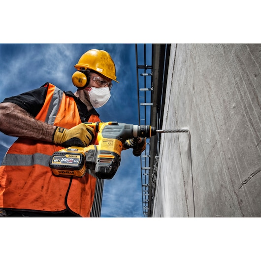 A construction worker wearing an orange safety vest, yellow gloves, and a yellow hard hat is using a DEWALT cordless power drill to bore into a concrete wall on a building site. The worker’s face is obscured.