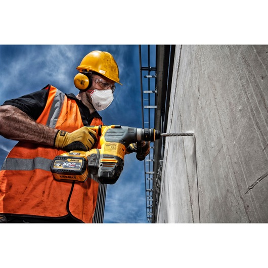 A construction worker wearing a yellow hard hat, orange safety vest, and gloves is using a DEWALT cordless rotary hammer drill (model DT9611) to drill into a concrete wall outdoors.