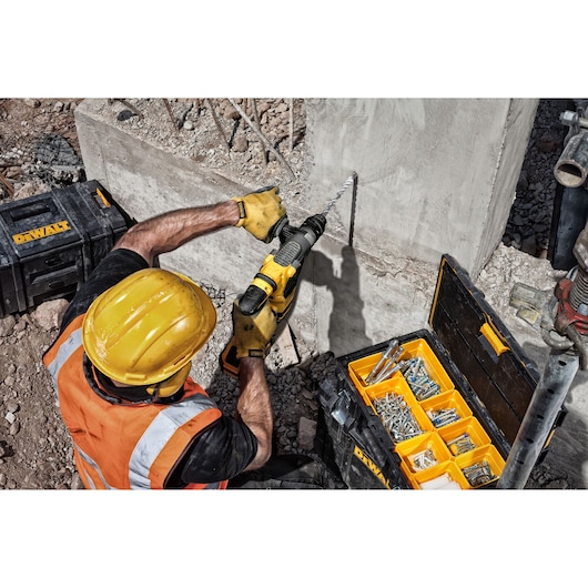 A construction worker wearing a yellow safety helmet and orange vest uses a DEWALT drill to bore a hole into a concrete wall. Nearby, a DEWALT black and yellow toolbox filled with screws and fasteners is open on the ground.
