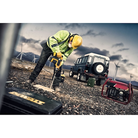 Worker using DEWALT drill on rocky ground near a vehicle, generator, and wind turbines, wearing safety gear.