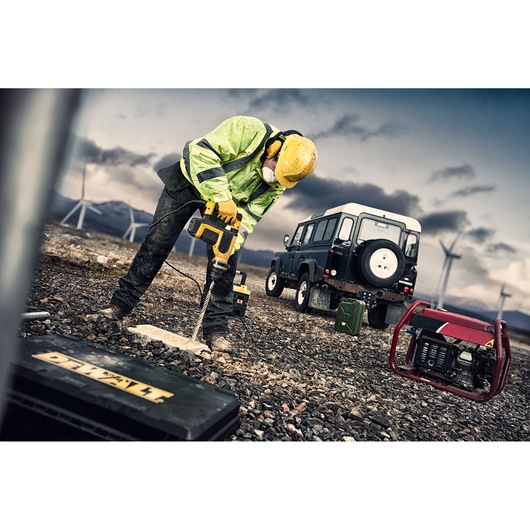 A construction worker wearing a yellow helmet and high-visibility jacket uses a DEWALT power tool to drill into the ground at an outdoor site. There is a black off-road vehicle and a generator nearby, with wind turbines and mountains visible in the background.