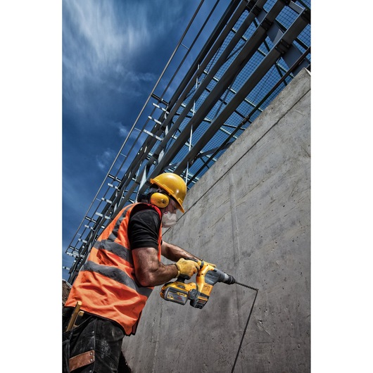 Worker in safety gear uses a DEWALT drill on a concrete wall at an outdoor construction site under blue sky.