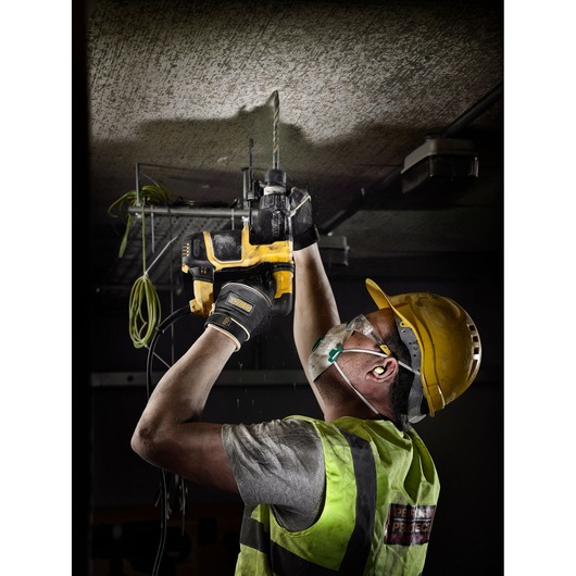 Worker wearing safety gear uses a DEWALT drill to work on a ceiling in a construction setting.