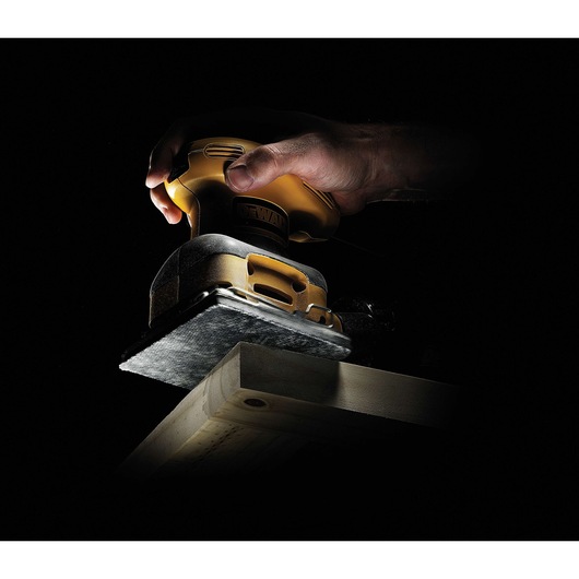 A hand using a DEWALT electric sander to smooth the surface of a wooden plank, with dramatic lighting highlighting the tool and wood against a dark background.