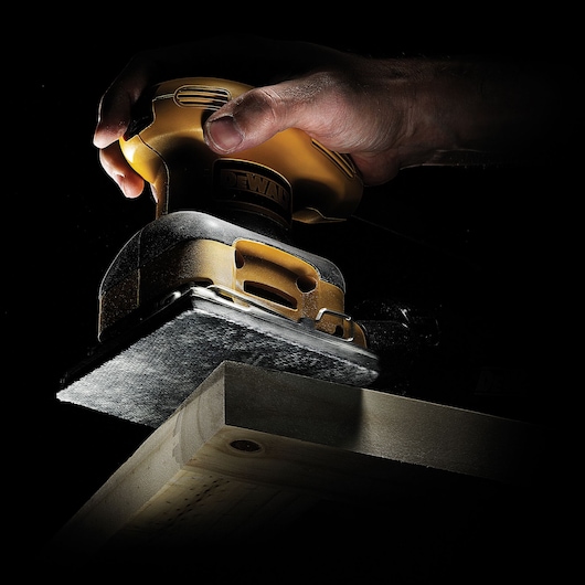 Hand using a DEWALT sander to smooth a wooden plank, close-up with dark background.