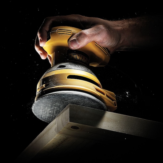 Hand holds DEWALT sander polishing wooden edge against dark background.