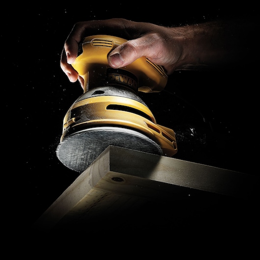 Hand holding a DEWALT sander, smoothing the edge of a wooden plank against a dark background.