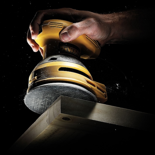 Hand using a DEWALT sander to smooth a wooden plank edge in a dark setting.
