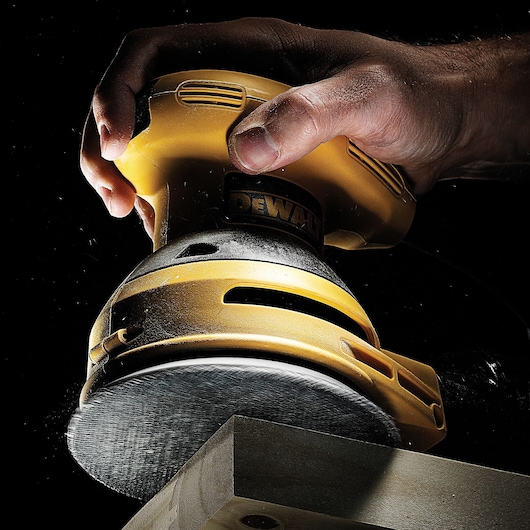 Hand holding DEWALT sander, smoothing wood surface. Close-up view, dust visible, black background.
