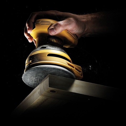 Hand using a DEWALT sander on a wooden plank, highlighting tool detail and sanding action.