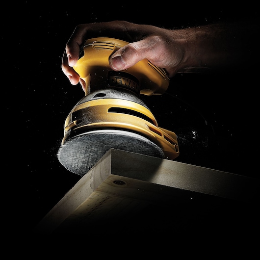 A close-up of a person using a yellow DEWALT electric sander to smooth the edge of a wooden plank, with dust particles visible against a black background.