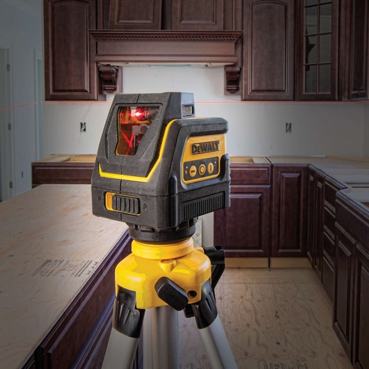 A DEWALT laser level device mounted on a yellow tripod is positioned in a kitchen under renovation, with visible cabinetry and unfinished countertops.