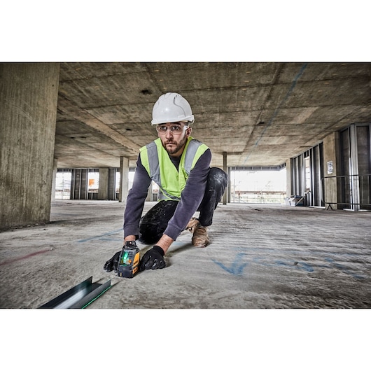 A worker wearing a hard hat, safety vest, and gloves is kneeling on a construction site floor and operating a DEWALT DW088CG green cross line laser tool for leveling and alignment.