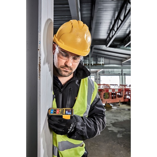 A person wearing a yellow hard hat, safety vest, and black gloves is using a DEWALT DW099S laser distance measurer against a vertical surface in an industrial indoor construction site.