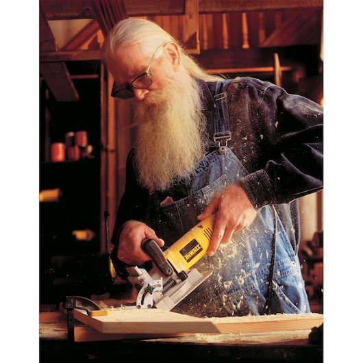 A person with a blurred face is using a DEWALT biscuit joiner (DW682K-QS) on a wooden board in a workshop. Sawdust is flying as the tool is in operation. The user is wearing denim overalls and a long-sleeve shirt.