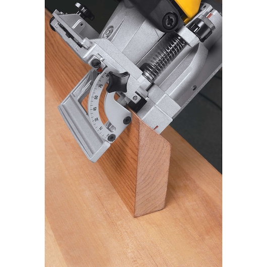 Close-up of a DEWALT electric biscuit joiner tool being used to cut a slot in a piece of wood at an angle. The tool shows a precise angle adjustment gauge and is positioned against a wooden surface.