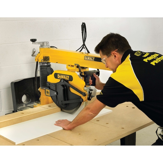 A person using a DEWALT radial arm saw to cut a piece of wood on a workbench. The saw is bright yellow and black, and the person's face is intentionally blurred.