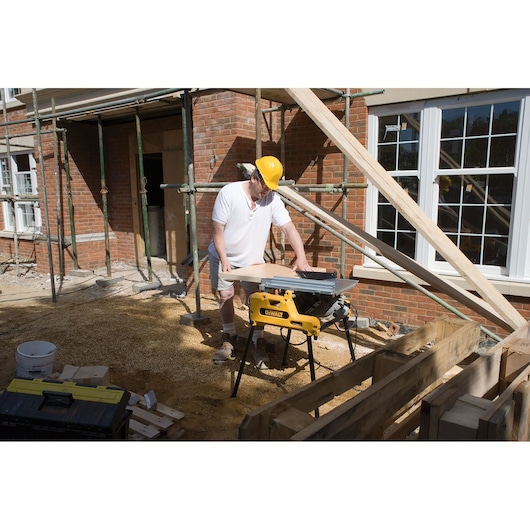 A construction worker wearing a yellow hard hat uses a DEWALT DW743N saw to cut wood at an outdoor building site, with scaffolding and a brick house in the background.