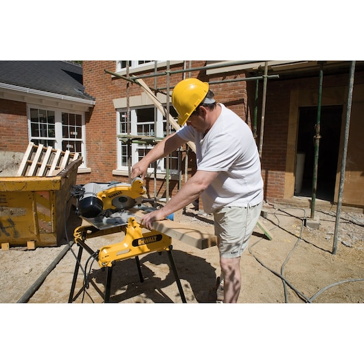 A person wearing a yellow hard hat uses a DEWALT DW743N saw to cut a piece of wood at a construction site, with scaffolding and a brick building in the background.