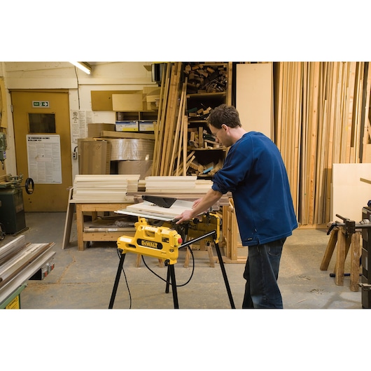 A person using a yellow DEWALT DW743N saw in a woodworking workshop, surrounded by stacked wood and various carpentry tools.