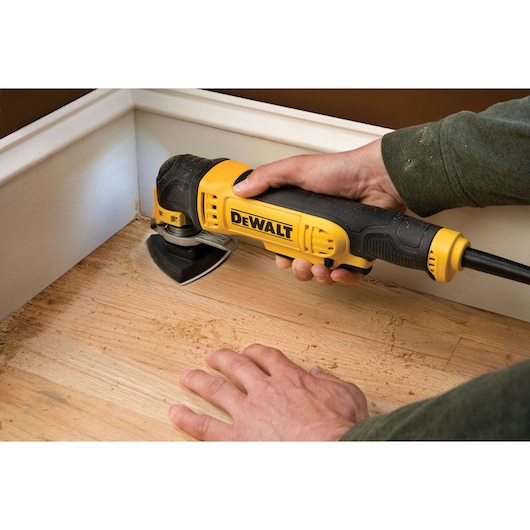A person using a DEWALT DWE315KT multi-tool to sand a wooden floor near a baseboard.