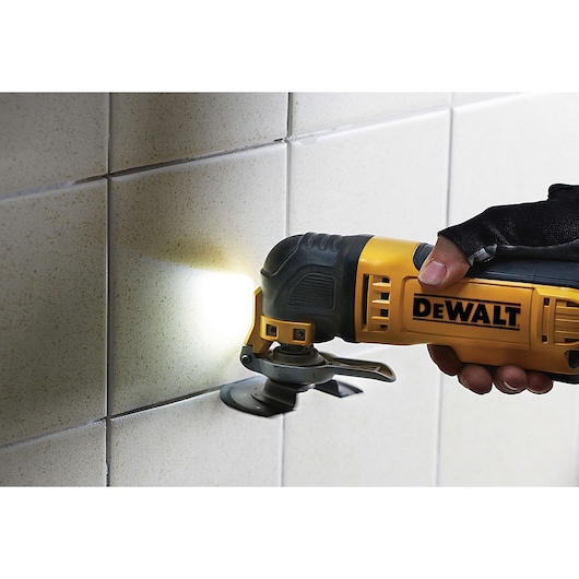 A person is using a DEWALT oscillating tool to work on the grout lines of wall tiles, with a light illuminating the area.