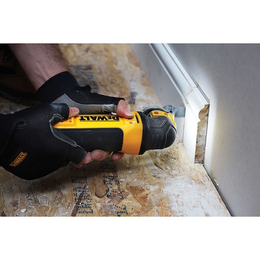A person wearing gloves uses a DEWALT multi-tool to cut into a wall beside a floor, with the tool illuminated for visibility.