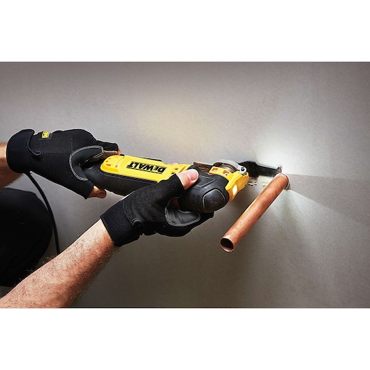 A person wearing black gloves is using a DEWALT oscillating multi-tool to cut drywall around a copper pipe.