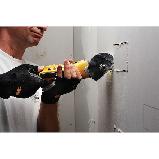 A person wearing black gloves is using a yellow DEWALT multi-tool to cut a rectangular opening in a drywall panel.