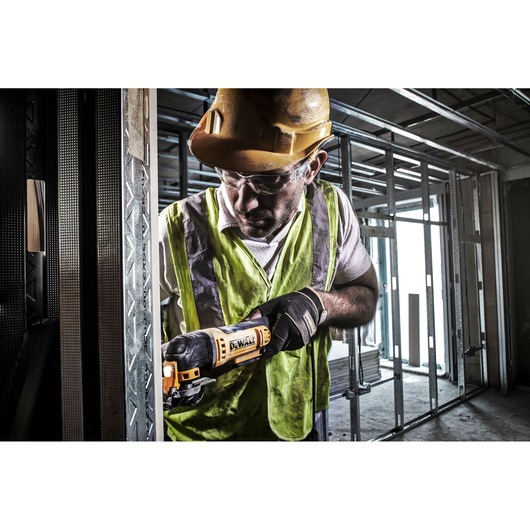 A construction worker wearing a safety vest and hard hat uses a DEWALT DWE315 oscillating tool to cut metal framing inside a building under construction. The worker's face is blurred for privacy.
