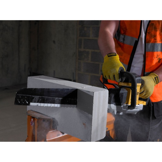 A person wearing yellow gloves and an orange safety vest is using a DEWALT power saw to cut through a grey concrete block indoors. Dust is visible around the area being cut.