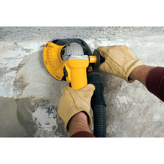 Close-up of gloved hands using a yellow DEWALT angle grinder with dust shroud attachment to smooth a concrete surface.