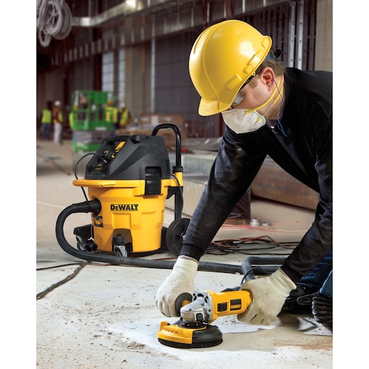 A construction worker wearing a yellow hard hat, gloves, and a face mask is using a DEWALT angle grinder with a dust shroud on a concrete floor. A DEWALT vacuum is connected to the grinder, helping to collect dust. The scene is set inside an unfinished building with other workers visible in the background.