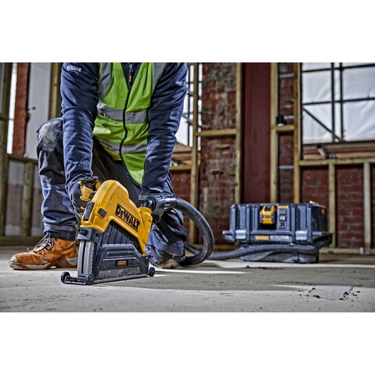 A person wearing work boots and a safety vest is using a DEWALT power tool to cut into a concrete floor, with a connected hose for dust extraction. In the background, there is a DEWALT storage box in a construction site setting.