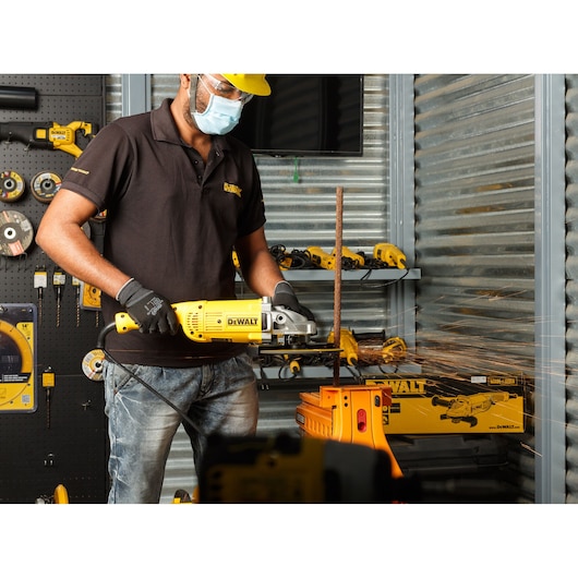 A person wearing gloves and a black DEWALT polo shirt is using a yellow DEWALT angle grinder (model DWE492) to cut a metal rod in a workshop. Sparks are visible as the tool operates. The background features various DEWALT tools and accessories organized on shelves and walls.