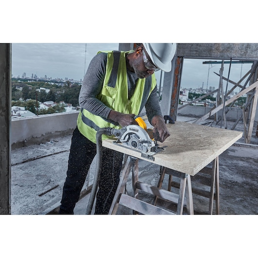 A construction worker wearing a safety vest and helmet is using a DEWALT DWE5615 circular saw to cut a wooden board on a building site.