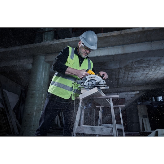 A person wearing a safety helmet and reflective vest is using a DEWALT DWE5615 circular saw to cut wood on a construction site. The background shows a partially constructed building and scattered construction materials.