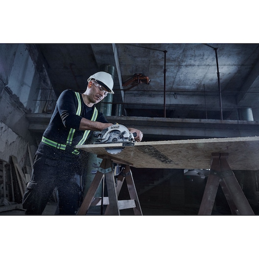 A person wearing a safety helmet and reflective vest is using a DEWALT circular saw to cut a wooden board supported by two sawhorses in a construction environment.