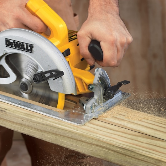 Close-up of a person using a DEWALT DWE575K circular saw to cut a wooden plank, with visible wood dust and motion blur from the spinning blade.
