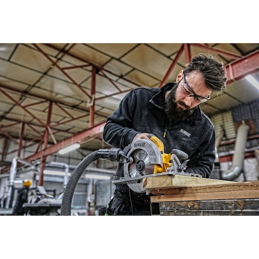 A person using a DEWALT circular saw to cut a piece of wood in a workshop. The person's face is blurred for privacy.