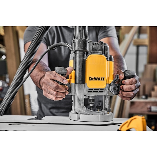 A person operating a DEWALT DWE625KT plunge router, with hands on the handles, in a woodworking environment.