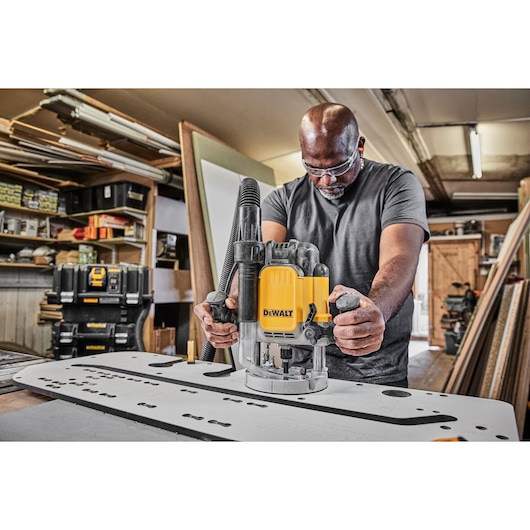 A person using a DEWALT DWE625KT router in a woodworking workshop, guiding the tool along a flat surface with a dust extraction hose attached. Various DEWALT storage cases and woodworking tools are visible in the background.
