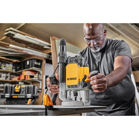 Person using a DEWALT router tool (model DWE625KT) on a wooden surface in a workshop environment. The tool is yellow and black, and there are various equipment and storage boxes visible in the background.
