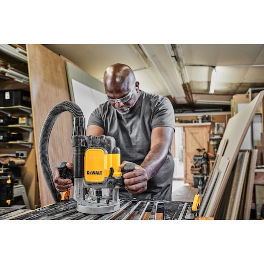 A person using a DEWALT DWE625 router tool in a woodworking workshop. The tool is placed on a workbench with wood pieces and a vacuum hose attached. The workshop is filled with various materials and equipment.