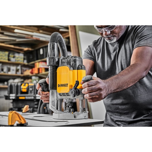 A person using a DEWALT DWE625 router on a workbench in a workshop. The yellow and black tool is equipped with a dust extraction hose. Other DEWALT tools and shelves with supplies are visible in the background.
