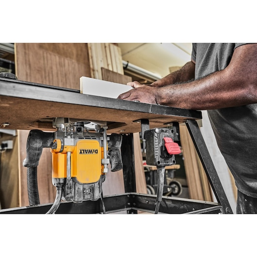 A DEWALT router mounted under a worktable in a workshop, with a person guiding a piece of wood along the table for woodworking.